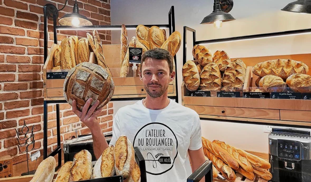 Un artisan boulanger présente une miche de pain ronde devant des étagères de pains variés.