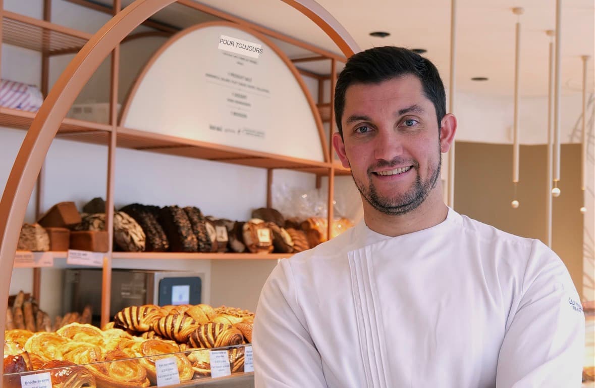 Boulanger souriant en veste blanche devant un étalage de pains et de viennoiseries artisanales.